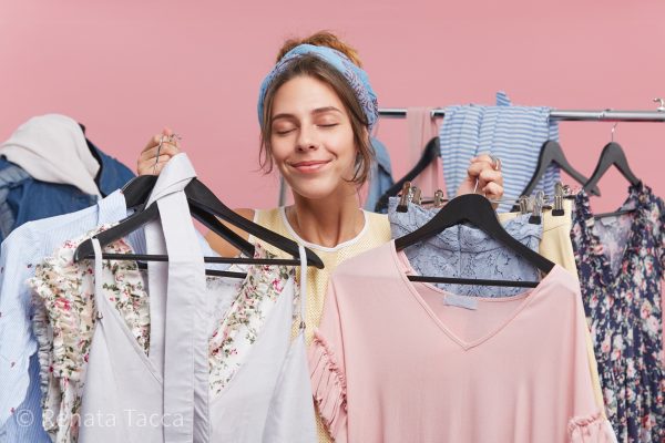 Pleased female model closing her eyes with pleasure while standing in cloak room, holding many hangers with clothes wanting to buy everything. Female shopaholic trying new clothes, refreshing wardrobe